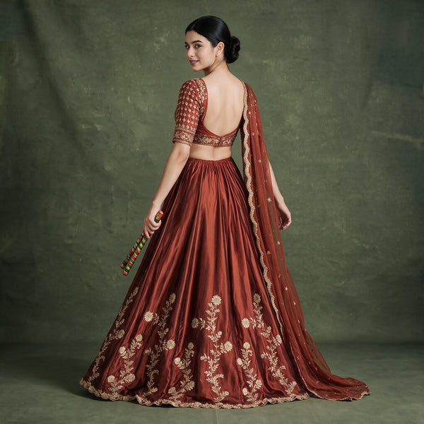 Back view of the rust lehenga choli with open-back blouse, gold floral embroidery on the skirt and the dupatta trailing behind