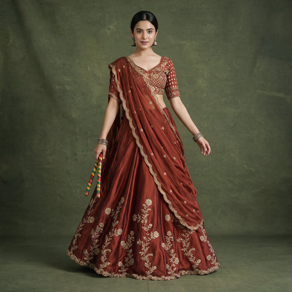 Model in a rust rangoli silk lehenga choli with gold floral embroidery on the skirt and a sheer rust dupatta with a scalloped gold edge, holding dandiya sticks