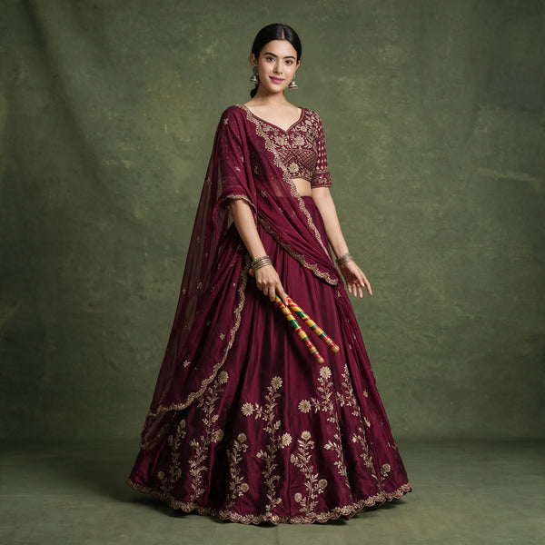 Model in a wine rangoli silk lehenga choli with gold thread embroidery on the skirt hem and a matching sequin-embroidered dupatta, holding dandiya sticks