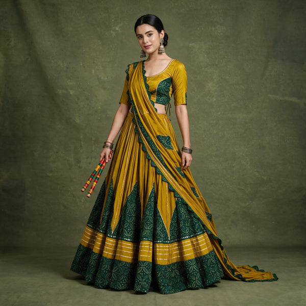 Model in a mustard rayon chaniya choli with green printed panels rising in points from the hem, a matching blouse and a mustard dupatta, holding dandiya sticks