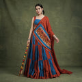 Model in a rust and blue panelled chaniya choli with a rust dupatta carrying a gamthi panel, holding dandiya sticks