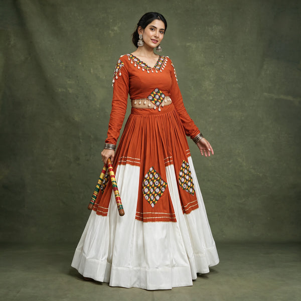 Model in an orange full-sleeve chaniya choli with a white tiered skirt, patchwork diamonds and cowrie shell trim, holding dandiya sticks