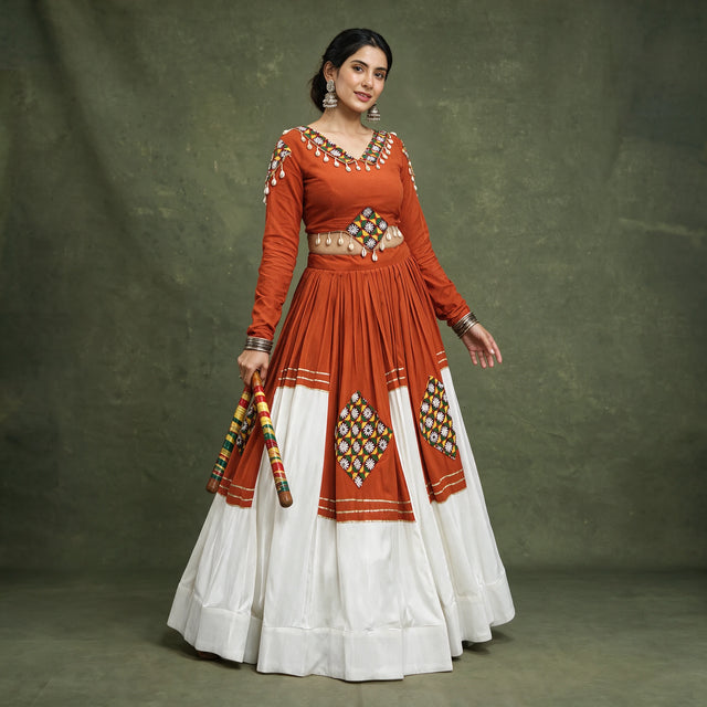 Model in an orange full-sleeve chaniya choli with a white tiered skirt, patchwork diamonds and cowrie shell trim, holding dandiya sticks