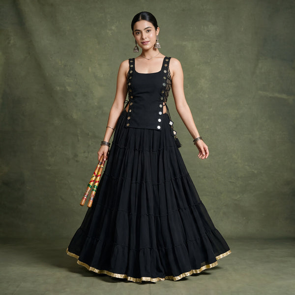 Model in a black tiered chaniya choli with a sleeveless koti studded with round silver discs and a gold-edged hem, holding dandiya sticks