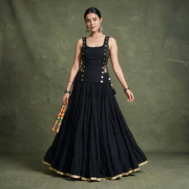 Model in a black tiered chaniya choli with a sleeveless koti studded with round silver discs and a gold-edged hem, holding dandiya sticks