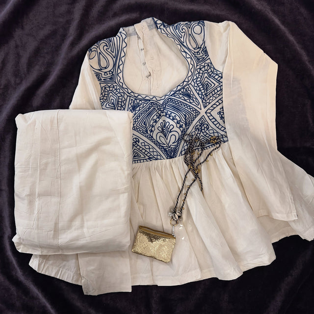 Flat lay of the ivory cotton top with a blue embroidered yoke, the ivory skirt and the small gold purse