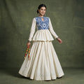 Model in an ivory cotton peplum top with a blue embroidered yoke over an ivory tiered skirt, holding dandiya sticks