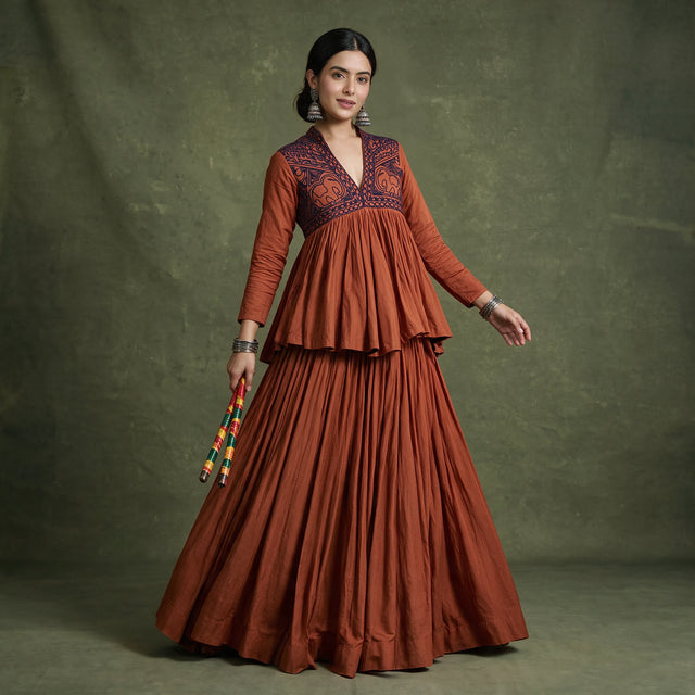 Model in a rust peplum top with a dark embroidered yoke over a full rust skirt, holding dandiya sticks