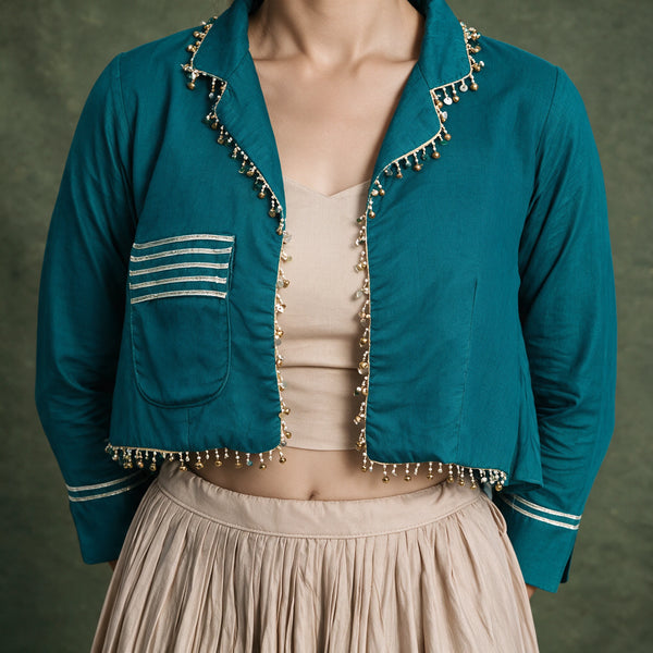 Close-up of the teal jacket with a striped pocket and gold bead edging over the beige top