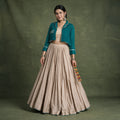 Model in a teal pure cotton short jacket with gold bead edging over a beige crop top and a pleated beige skirt, holding dandiya sticks