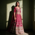Model in a raspberry raw silk bridal lehenga with gold leaf-vine columns, a tiered embroidered hem, V-neck blouse, net dupatta and beaded latkan in window light