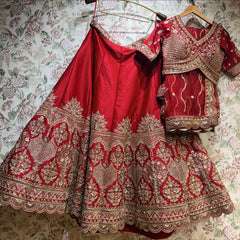 Red raw silk lehenga and cross-over blouse with net dupatta hung against floral wallpaper, showing the peacock-fan and floral-arch hem and scalloped border