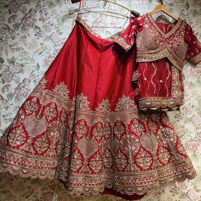 Red raw silk lehenga and cross-over blouse with net dupatta hung against floral wallpaper, showing the peacock-fan and floral-arch hem and scalloped border