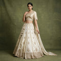 Model in an ivory silk lehenga with gold embroidered motifs on vertical panels, scalloped hem, embroidered blouse and draped dupatta