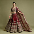 Model in a maroon raw silk bridal lehenga with gold cow motif hem border, embroidered V-neck blouse and maroon net dupatta against an olive backdrop