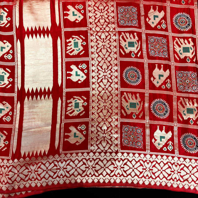 Red saree fabric with geometric squares, floral medallions, and patterned border trim.
