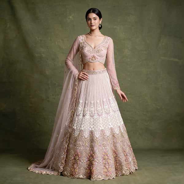 Model in a baby pink net lehenga with ivory pearl and cutdana embroidery fading into a gold and floral hem, sheer long-sleeve blouse and net dupatta