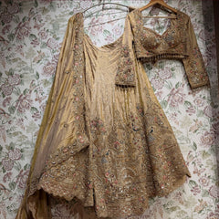 Antique gold tissue silk lehenga, embroidered blouse and cape dupatta hung against floral wallpaper, showing the peacock and floral hem and scalloped dupatta border