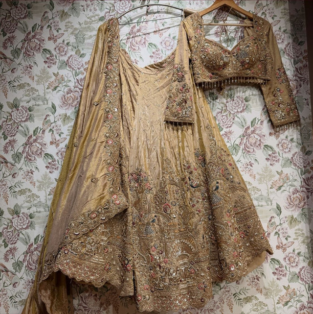 Antique gold tissue silk lehenga, embroidered blouse and cape dupatta hung against floral wallpaper, showing the peacock and floral hem and scalloped dupatta border