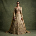 Model in an antique gold tissue silk lehenga with a peacock garden embroidered hem, full-sleeve sweetheart blouse with beaded fringe, long cape dupatta and beaded latkan, against an olive backdrop
