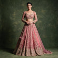 Model in a dusty rose pink lehenga with all-over gold cutdana and pearl embroidery, a leaf and floral hem, full-sleeve sweetheart blouse with beaded fringe, net dupatta and tassel latkan against an olive backdrop