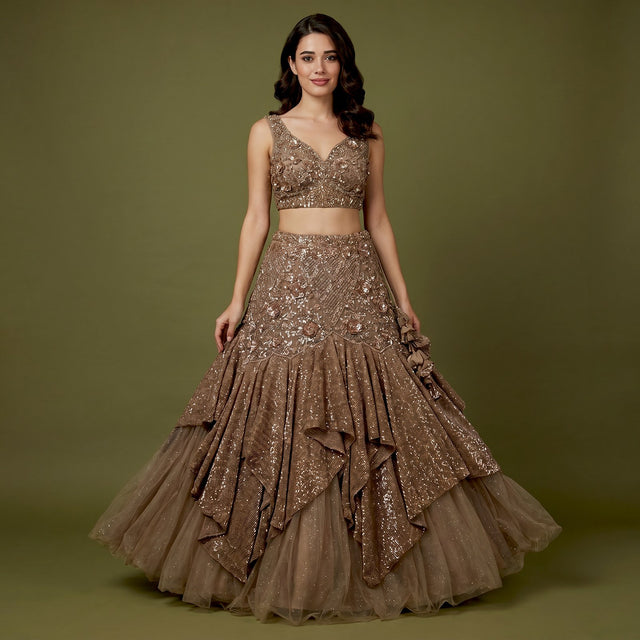 Model in a bronze brown net fish cut lehenga with sequinned ruffle tiers, a fitted 3D floral yoke and a sleeveless V-neck floral blouse against an olive backdrop