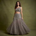 Model in a smoke grey net fish cut lehenga with a beaded asymmetric draped yoke and a sleeveless embroidered blouse with a flower at the shoulder against an olive backdrop