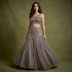 Model in a smoke grey net fish cut lehenga with a beaded asymmetric draped yoke and a sleeveless embroidered blouse with a flower at the shoulder against an olive backdrop