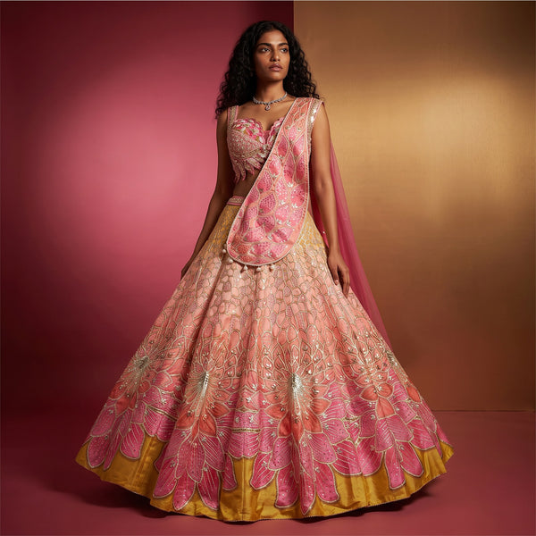Model in a peach-to-coral raw silk lehenga with pink and coral lotus-petal fans over a mustard hem, sequinned blouse and pink drape dupatta against a rose and gold backdrop