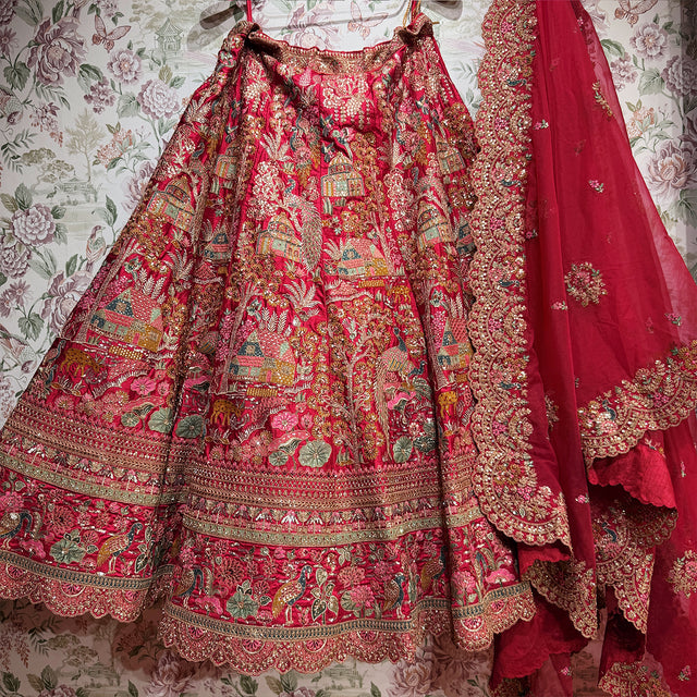 Pink raw silk lehenga skirt laid out showing peacocks, houses and trees above stacked zari and resham hem bands