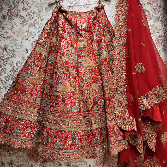 Red raw silk lehenga skirt laid out showing peacocks, houses and trees above stacked zari and resham hem bands