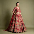 Model in a red raw silk bridal lehenga with village-scene meenakari resham embroidery, scalloped V-neck blouse and organza dupatta against an olive backdrop