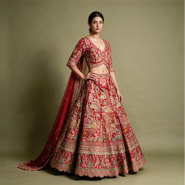 Model in a red raw silk bridal lehenga with village-scene meenakari resham embroidery, scalloped V-neck blouse and organza dupatta against an olive backdrop