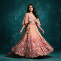 Model in a peach to coral ombre tissue silk lehenga with crane and floral embroidery at the hem, a poncho-style jacket with cape sleeves, a short blouse and a tasselled latkan against a teal backdrop