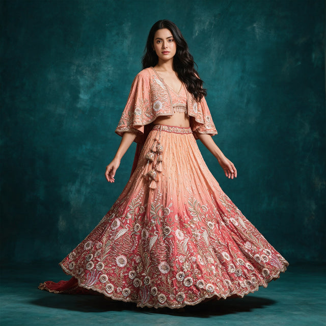 Model in a peach to coral ombre tissue silk lehenga with crane and floral embroidery at the hem, a poncho-style jacket with cape sleeves, a short blouse and a tasselled latkan against a teal backdrop