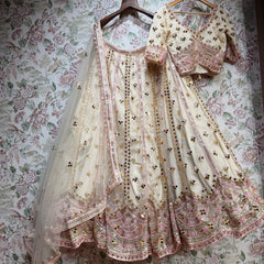 Ivory net lehenga and blouse on hangers against floral wallpaper, showing pink embroidery lines, mirror discs and the scalloped pink hem with a net dupatta