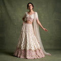 Model in an ivory net lehenga with vertical pink embroidery lines, a scalloped pink hem, cross-over V-neck blouse and sheer net dupatta against an olive backdrop