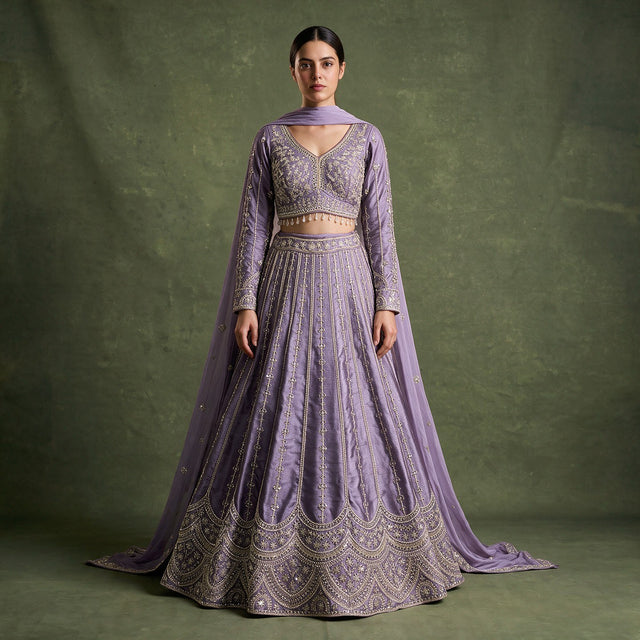Model in a lavender raw silk lehenga with slim embroidered floral lines, a scalloped arch hem, full-sleeve V-neck blouse with beaded drops and a matching dupatta against an olive backdrop