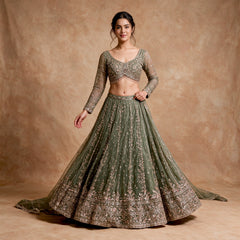 Model in an olive green net lehenga with all-over cutdana and sequin embroidery, a floral scalloped hem, full-sleeve blouse with beaded fringe and a net dupatta against a warm backdrop