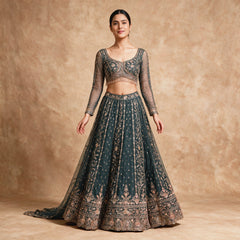 Model in a teal net lehenga with all-over cutdana and sequin embroidery, a floral hem, full-sleeve sheer blouse with beaded fringe and a net dupatta against a warm backdrop