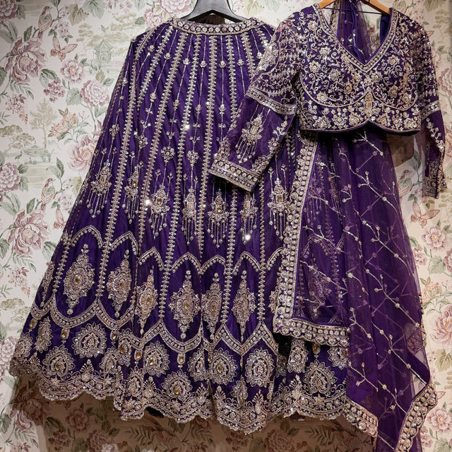 Purple net lehenga, embroidered full-sleeve blouse and purple net dupatta with a gold lattice on hangers against floral wallpaper