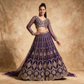 Model in a purple net lehenga with vertical bead lines, stone-set gold medallions and a scalloped hem, a full-sleeve V-neck blouse and a purple net dupatta against a warm gold backdrop