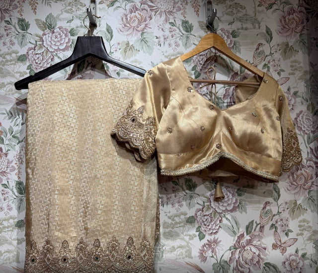 Folded champagne gold saree with beaded scalloped hem border beside the stitched gold blouse with floral motifs and embroidered sleeve cuffs, on hangers