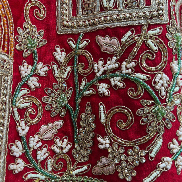 Red lehenga fabric with gold thread swirls, green floral accents, and white beaded motifs