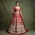 Model in a red bridal lehenga with peacock and arch handwork, hem bands of pearls and zari, sweetheart-neck blouse and net dupatta against a green backdrop