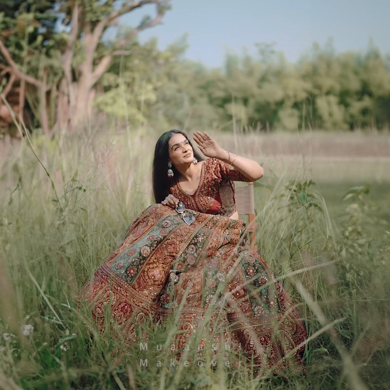 Elegant woman wearing a velvet maroon bridal lehenga in a lush outdoor setting with intricate designs